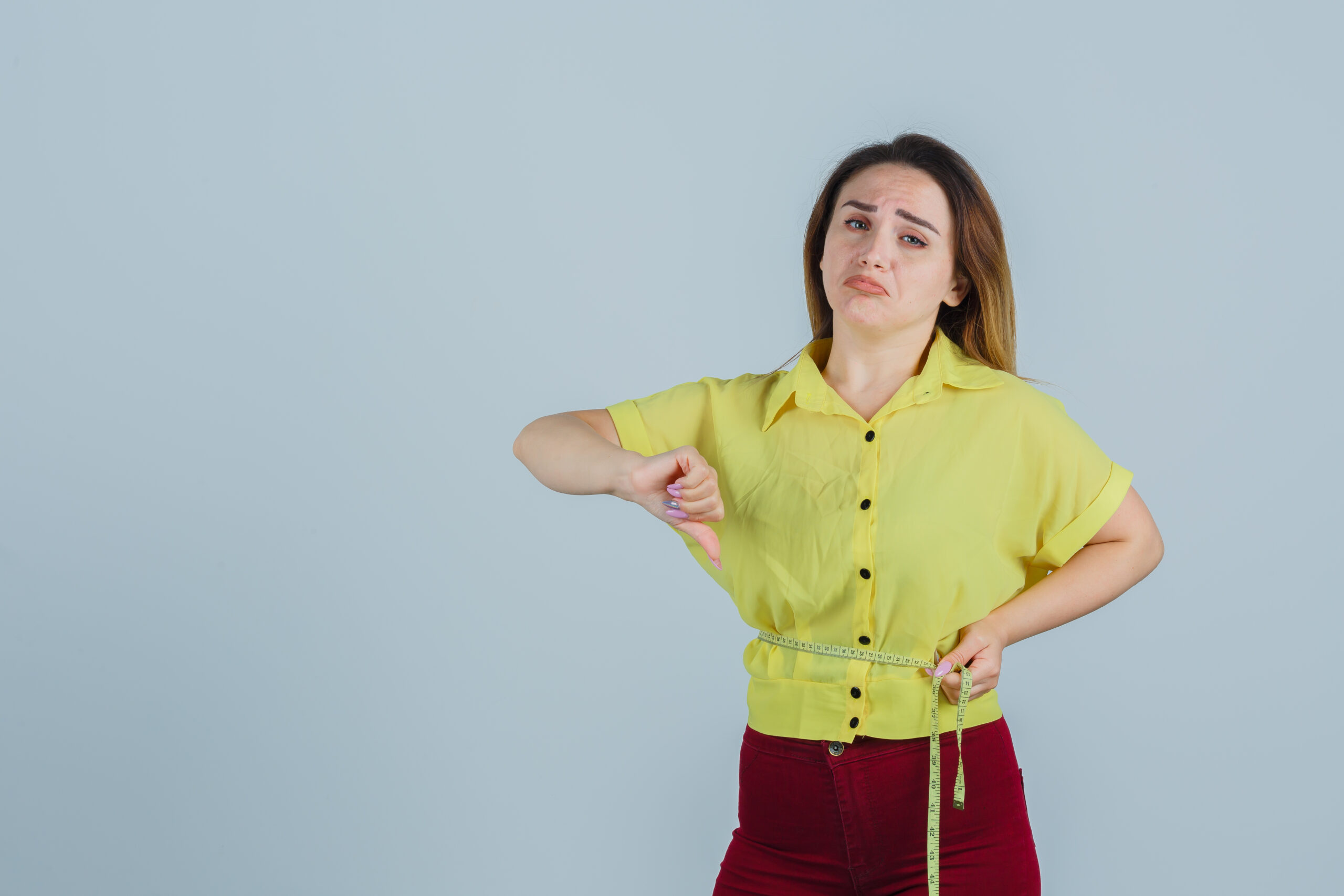 Disappointed woman measuring waist with tape measure and giving a thumbs-down sign.